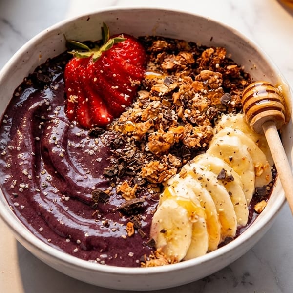 A vibrant acai bowl topped with granola, sliced bananas, a strawberry, and a honey dipper.