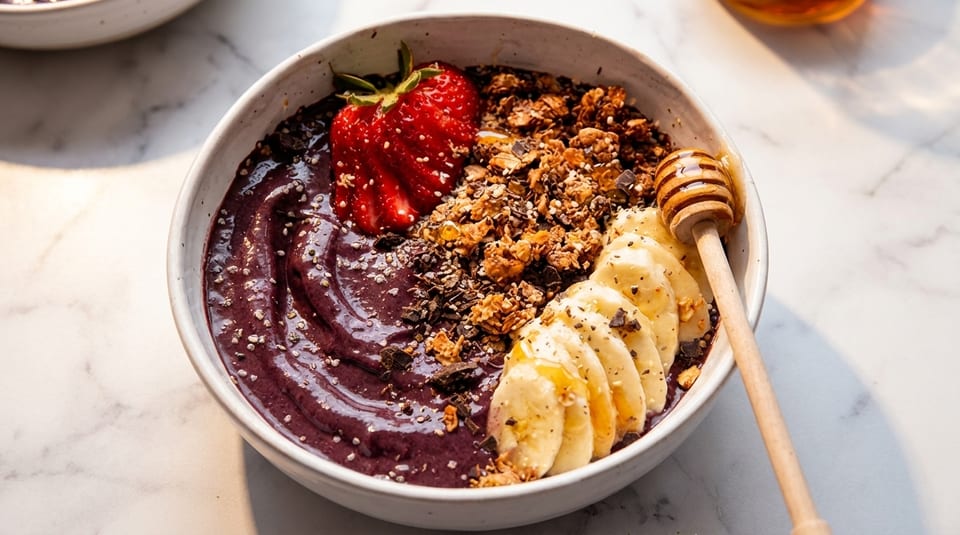 A vibrant acai bowl topped with granola, sliced bananas, a strawberry, and a honey dipper.