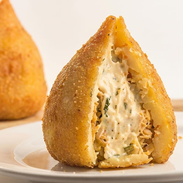 A golden-fried coxinha, cut open to reveal creamy chicken and herb filling, on a white plate.