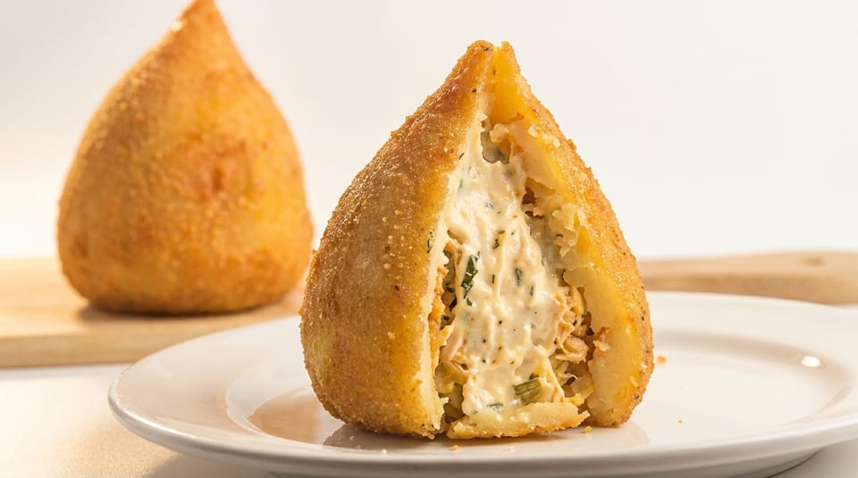 A golden-fried coxinha, cut open to reveal creamy chicken and herb filling, on a white plate.